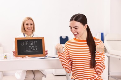 Photo of Happy woman lost weight and nutritionist with small blackboard in clinic, selective focus