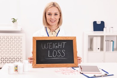 Photo of Happy nutritionist holding small blackboard with words Weight loss at table in clinic