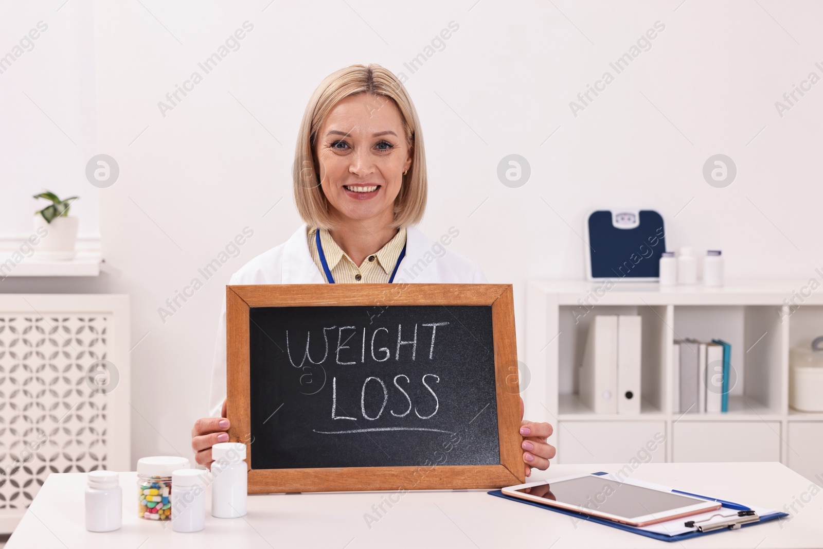 Photo of Happy nutritionist holding small blackboard with words Weight loss at table in clinic