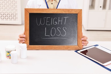 Photo of Nutritionist holding small blackboard with words Weight loss at table in clinic, closeup