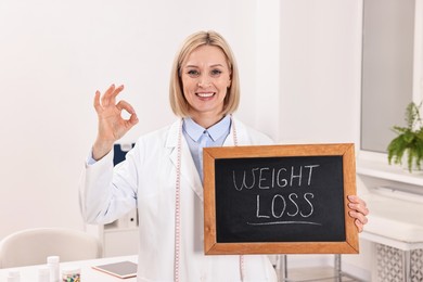 Photo of Happy nutritionist holding small blackboard with words Weight loss and showing ok gesture in clinic