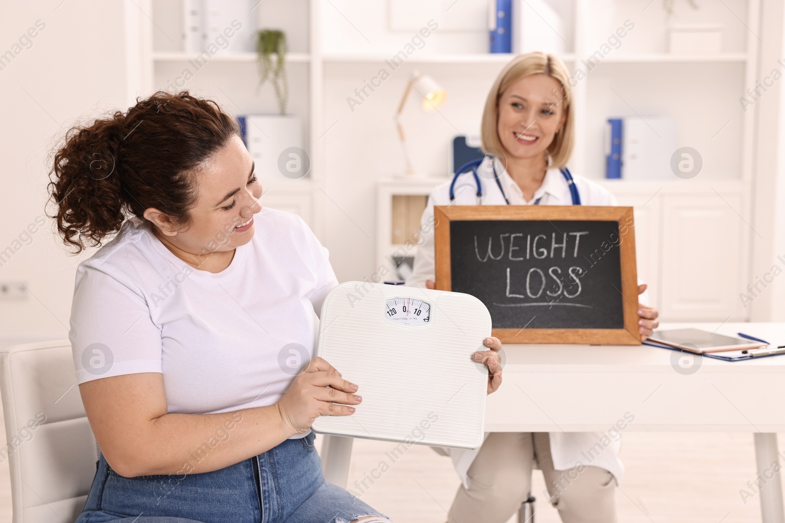 Photo of Happy woman with scales and nutritionist holding small blackboard with words Weight loss in clinic