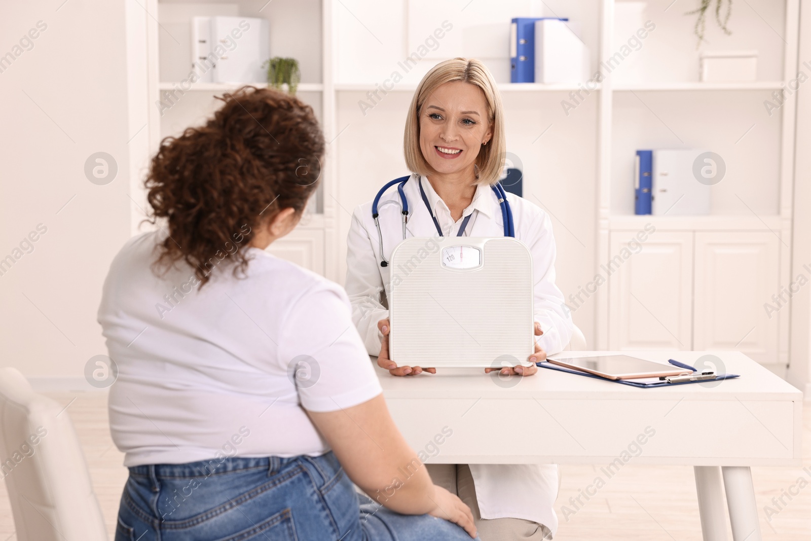 Photo of Weight loss. Smiling nutritionist with scales consulting patient at table in clinic