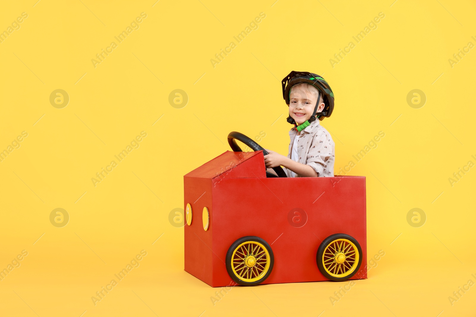 Photo of Little boy driving car made of cardboard on yellow background. Space for text