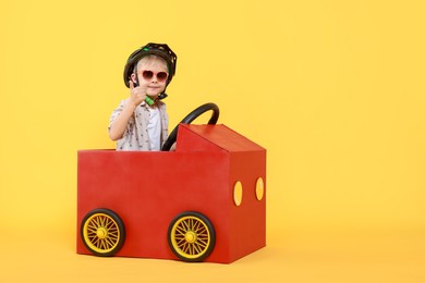 Photo of Little boy showing thumbs up while driving car made of cardboard on yellow background. Space for text