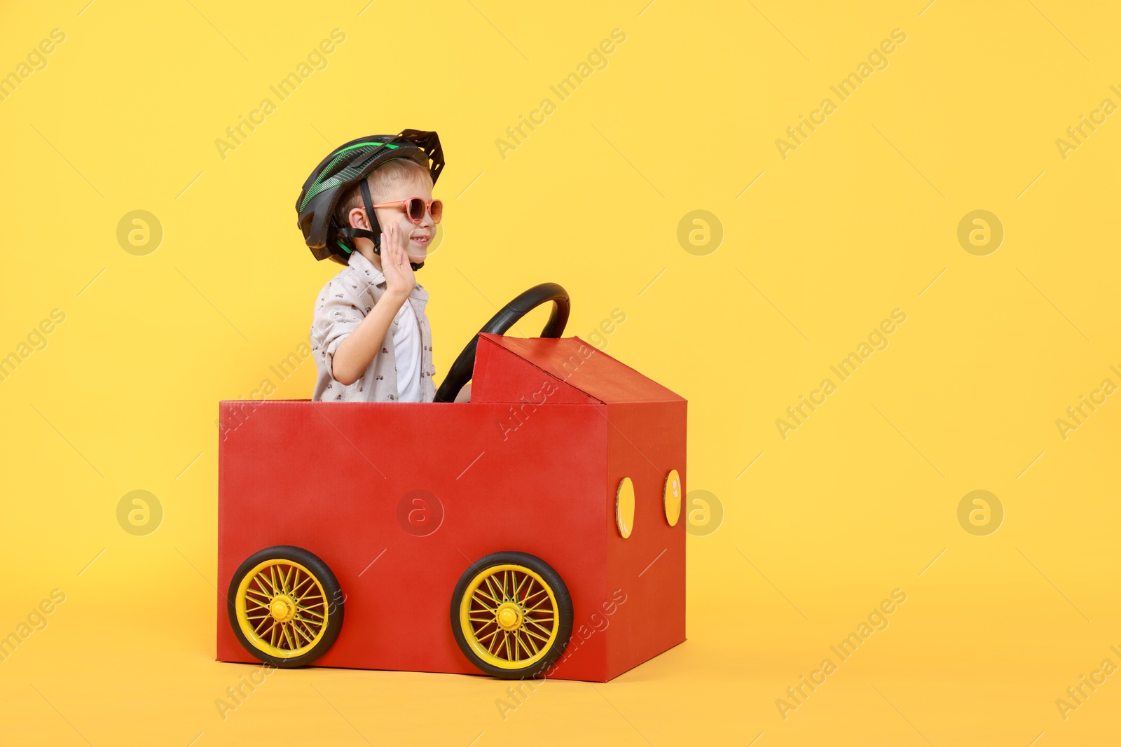 Photo of Little boy waving while driving car made of cardboard on yellow background. Space for text