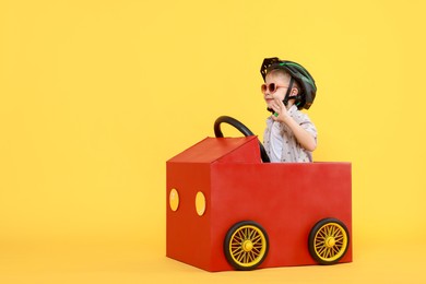 Photo of Little boy waving while driving car made of cardboard on yellow background. Space for text