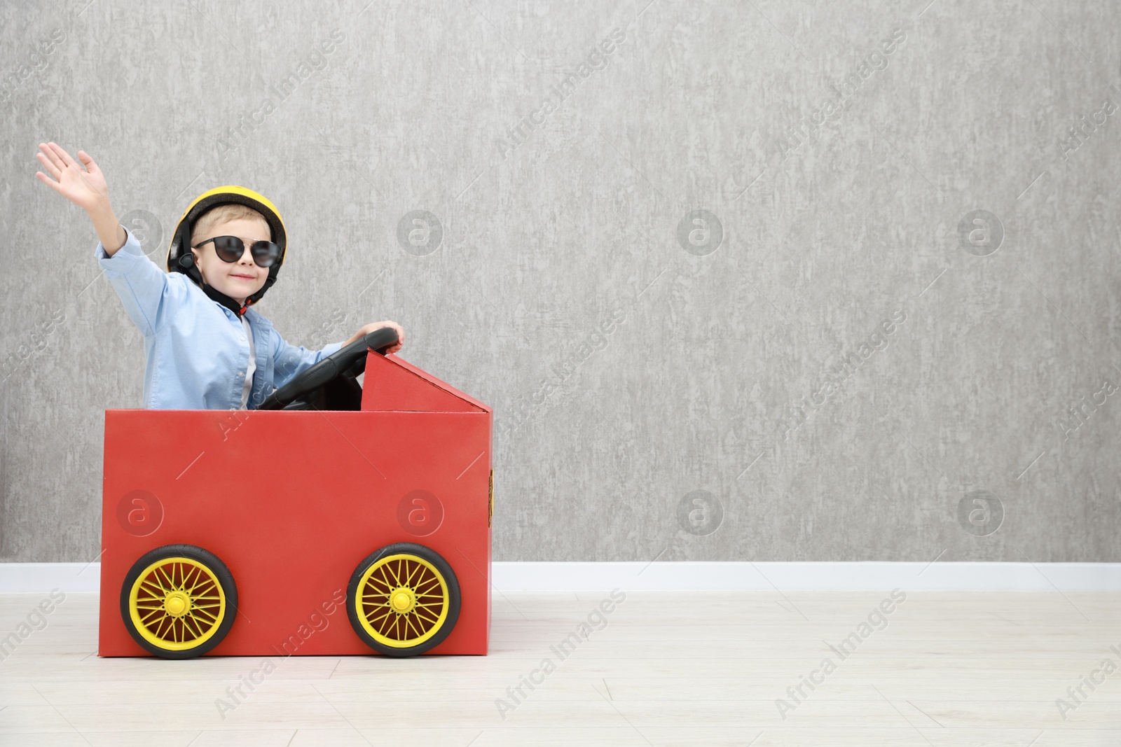 Photo of Little boy waving while driving car made of cardboard against grey wall. Space for text