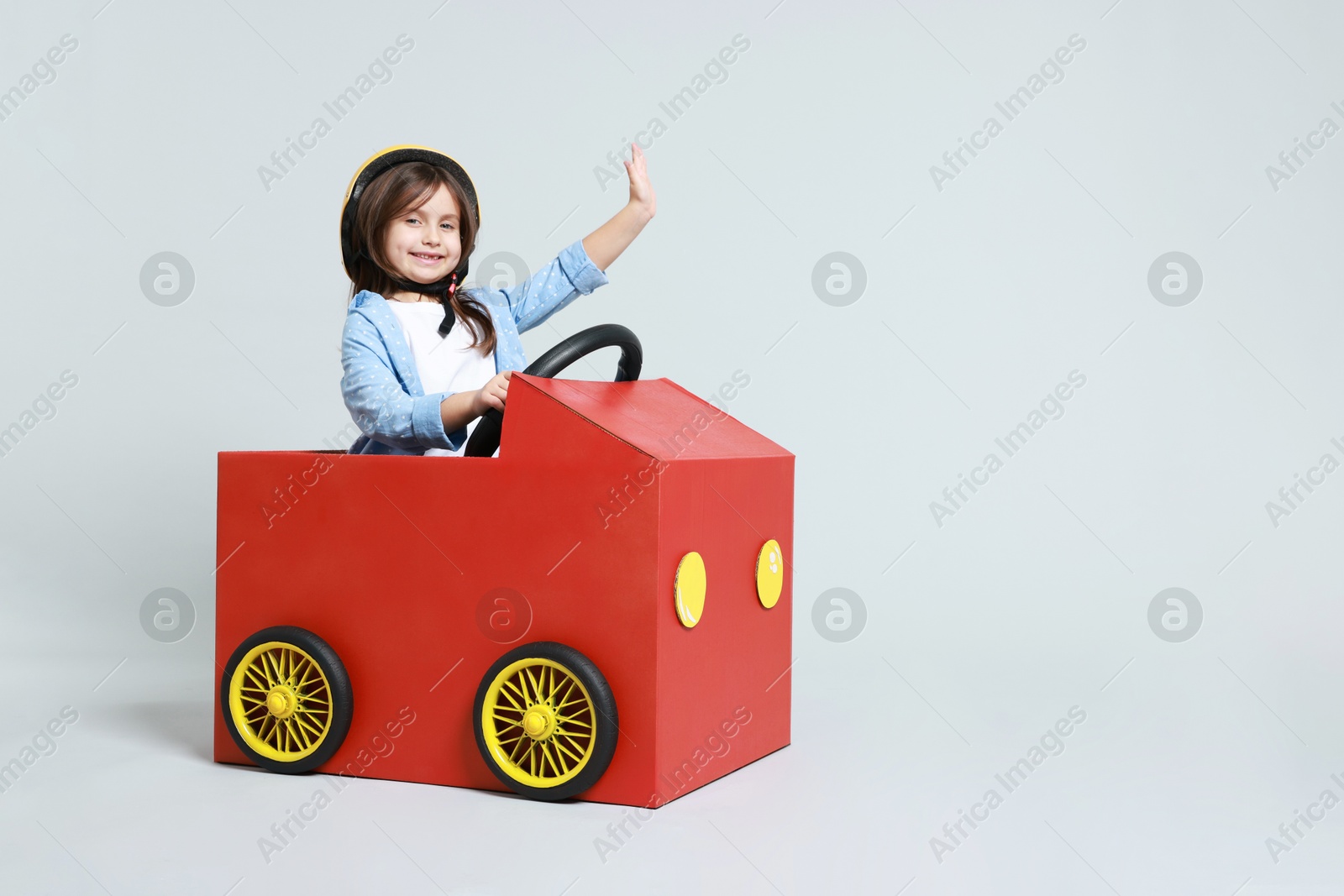 Photo of Little girl waving while driving car made with cardboard on light background. Space for text