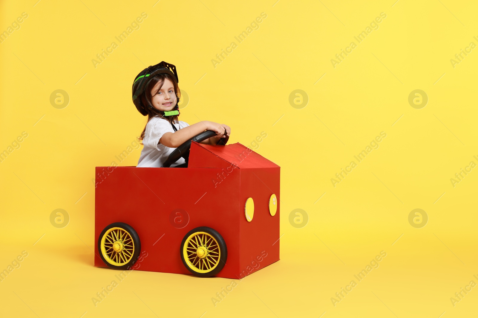 Photo of Little girl driving car made with cardboard on yellow background. Space for text