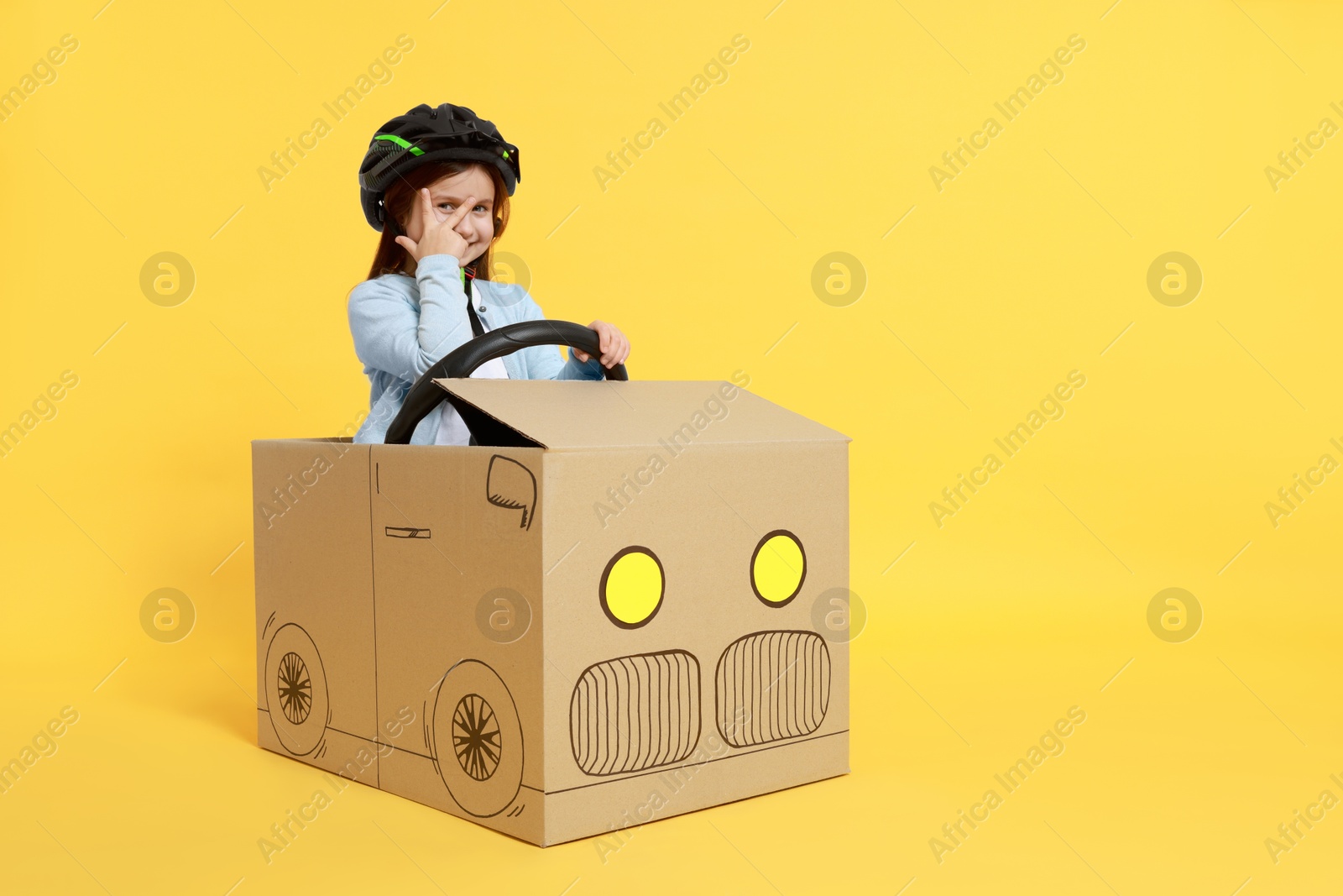 Photo of Little girl showing V-sign while driving car made with cardboard on yellow background. Space for text