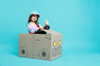 Photo of Little girl waving while driving car made with cardboard on light blue background. Space for text