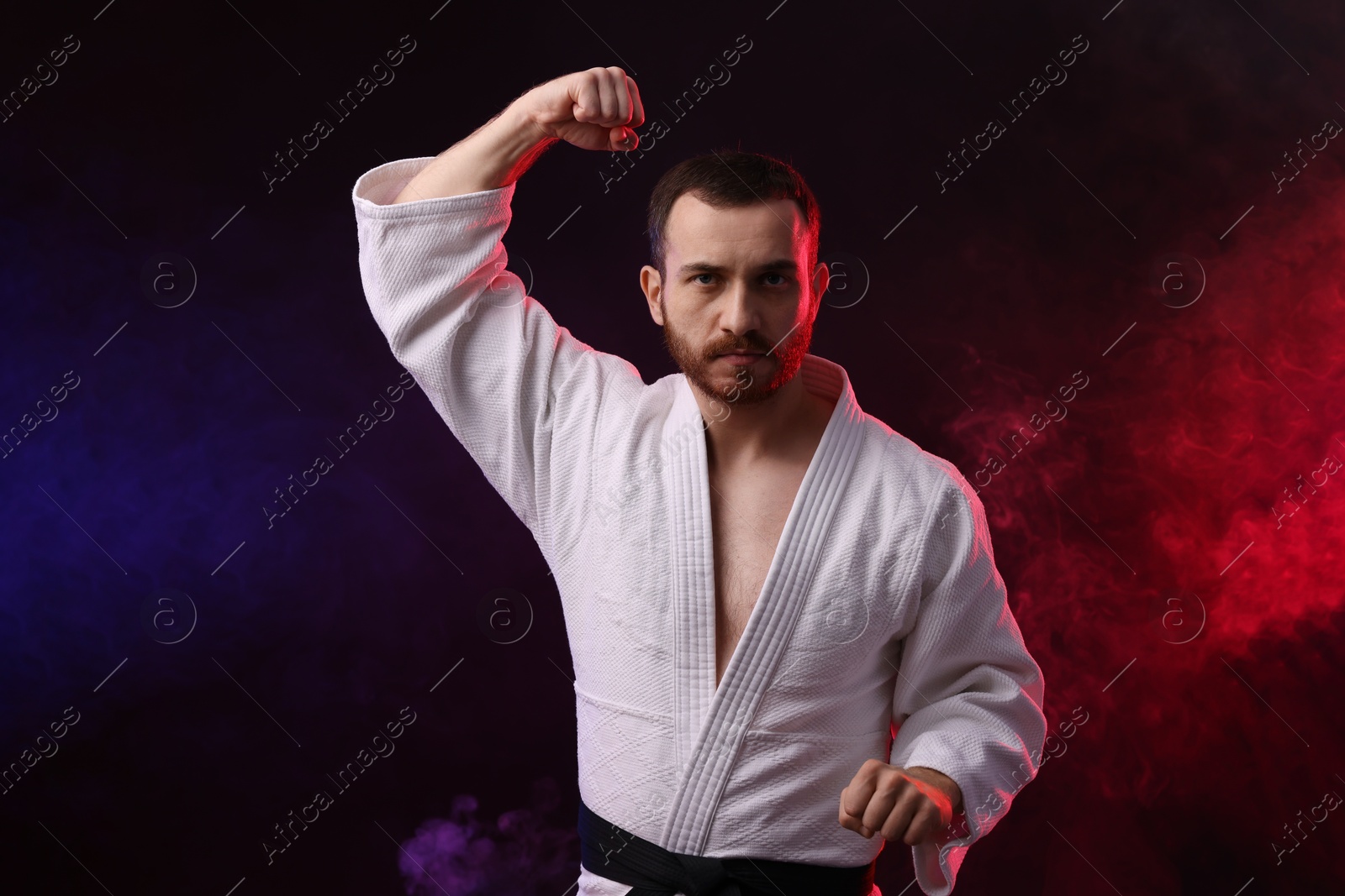 Photo of Man wearing uniform practicing karate in color lights and smoke against black background