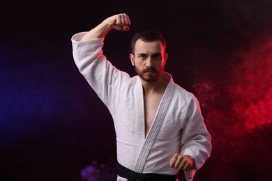 Photo of Man wearing uniform practicing karate in color lights and smoke against black background