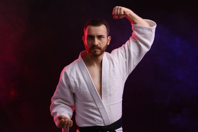 Photo of Man wearing uniform practicing karate in color lights and smoke against black background