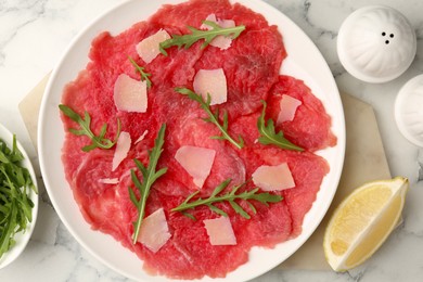 Delicious beef carpaccio with arugula, cheese and lemon on white marble table, flat lay