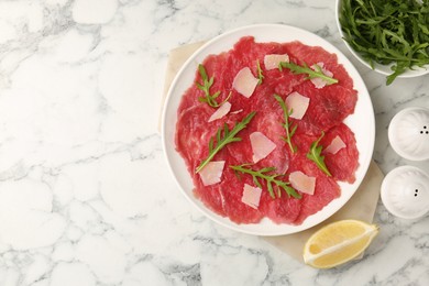 Delicious beef carpaccio with arugula, cheese and lemon on white marble table, flat lay. Space for text