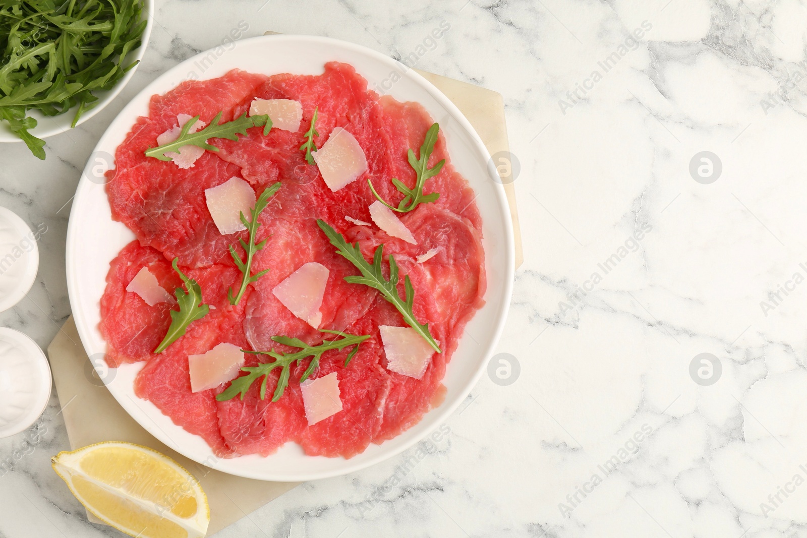 Photo of Delicious beef carpaccio with arugula, cheese and lemon on white marble table, flat lay. Space for text