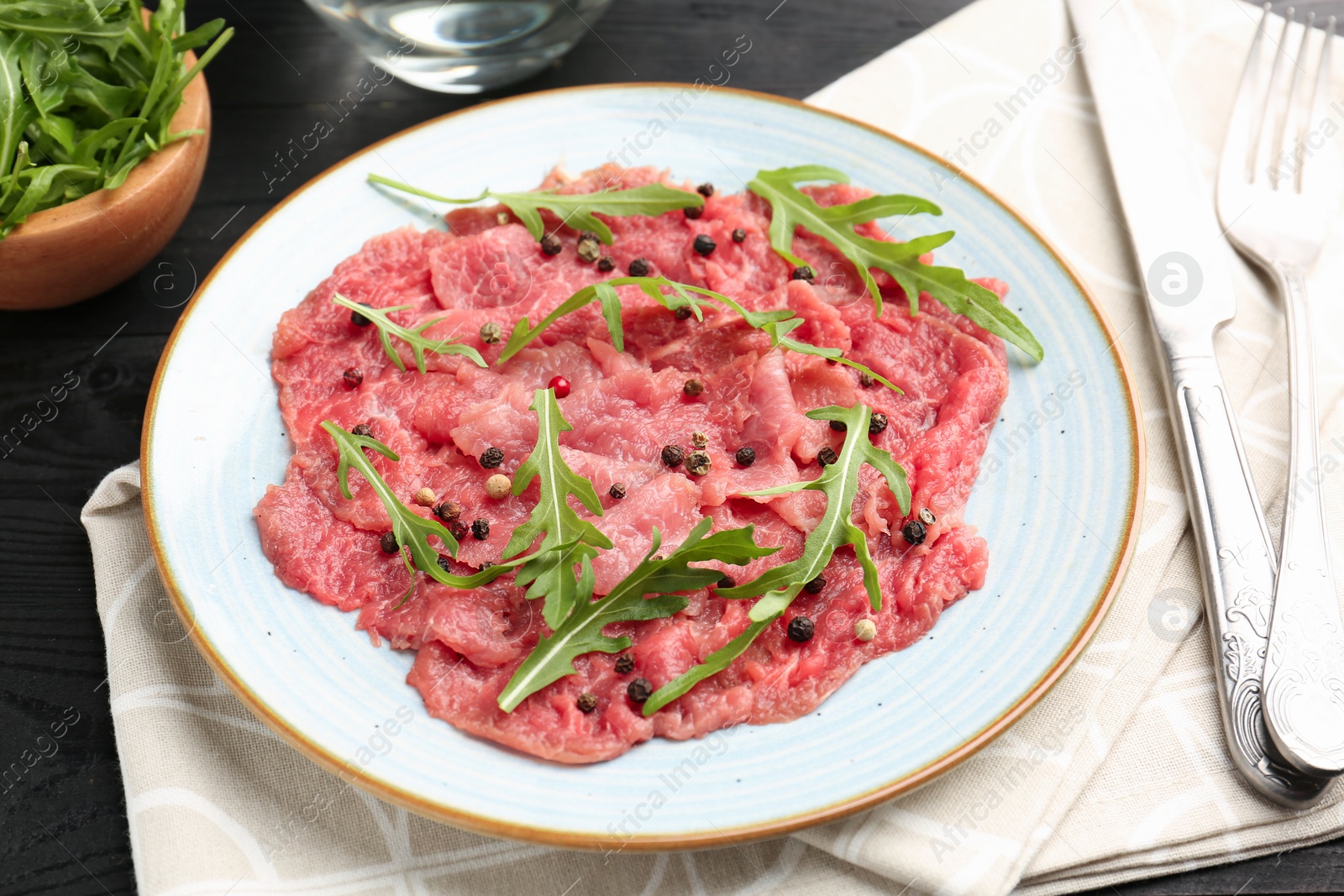 Photo of Fresh beef carpaccio with arugula and peppercorns on black wooden table, closeup