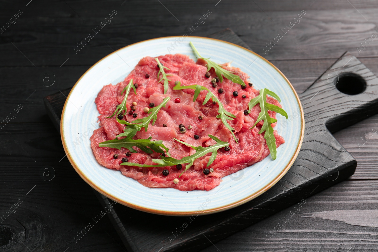 Photo of Fresh beef carpaccio with arugula and peppercorns on black wooden table, closeup