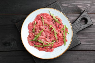 Photo of Fresh beef carpaccio with arugula and peppercorns on black wooden table, top view