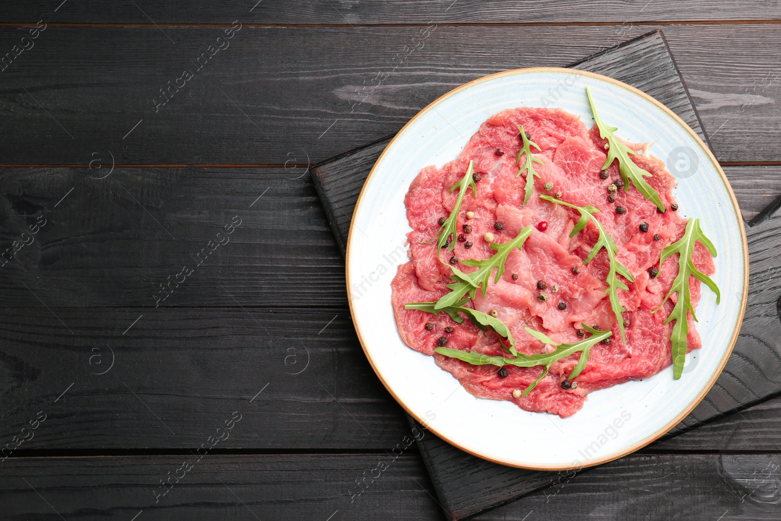 Photo of Fresh beef carpaccio with arugula and peppercorns on black wooden table, top view. Space for text