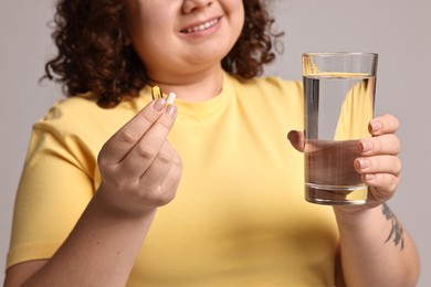 Photo of Happy plus size woman with weight loss supplements and glass of water on grey background, closeup