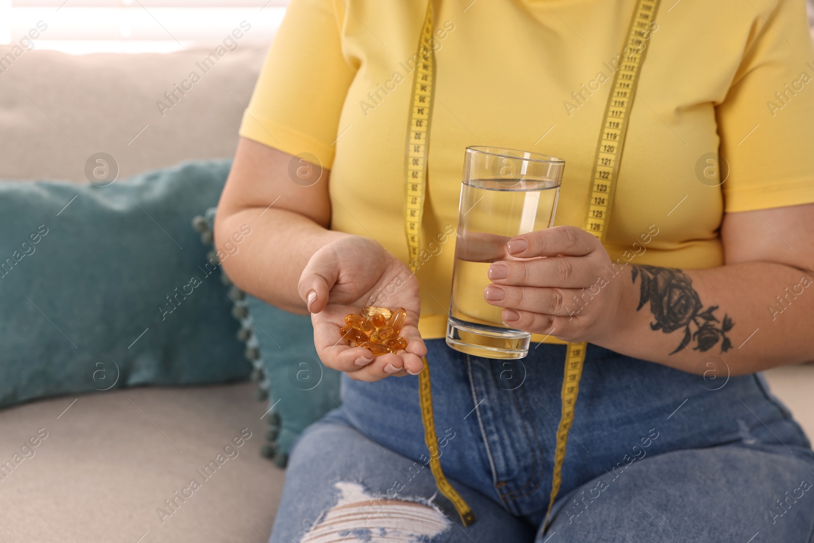 Photo of Plus size woman with pile of weight loss supplements and glass of water on sofa at home, closeup. Space for text