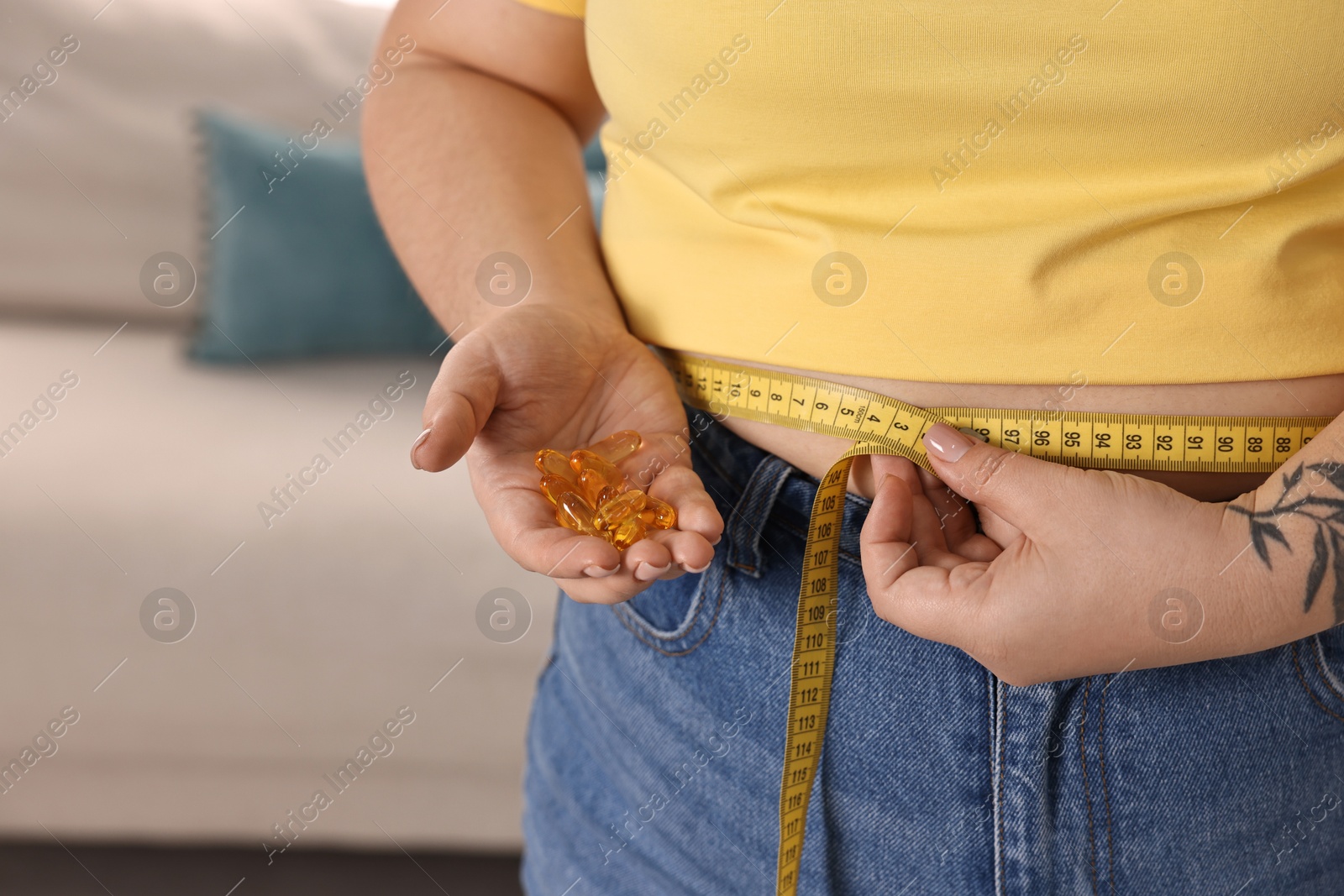 Photo of Plus size woman measuring waist with tape and holding pile of weight loss supplements at home, closeup. Space for text