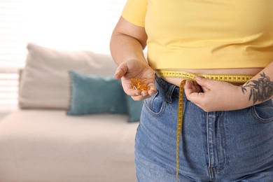 Photo of Plus size woman measuring waist with tape and holding pile of weight loss supplements at home, closeup. Space for text