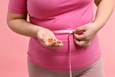 Photo of Plus size woman measuring waist with tape and holding pile of weight loss supplements on pink background, closeup