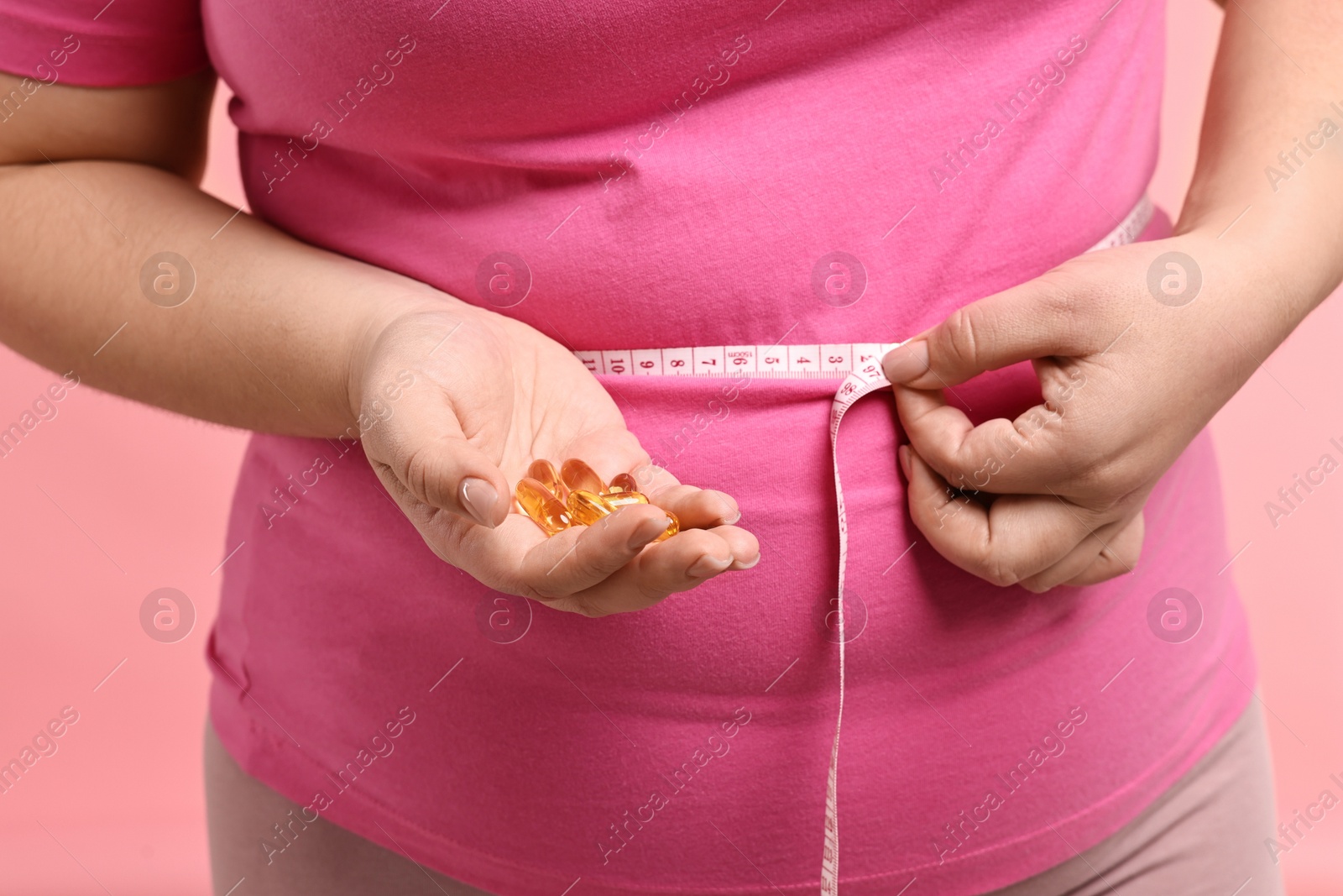 Photo of Plus size woman measuring waist with tape and holding pile of weight loss supplements on pink background, closeup