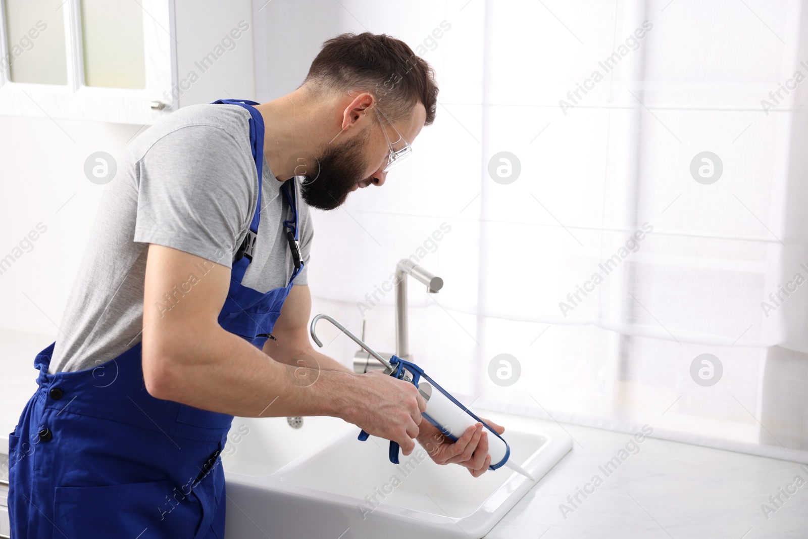 Photo of Worker with caulking gun sealing kitchen sink indoors