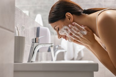 Photo of Woman washing her face with cleansing foam over sink in bathroom