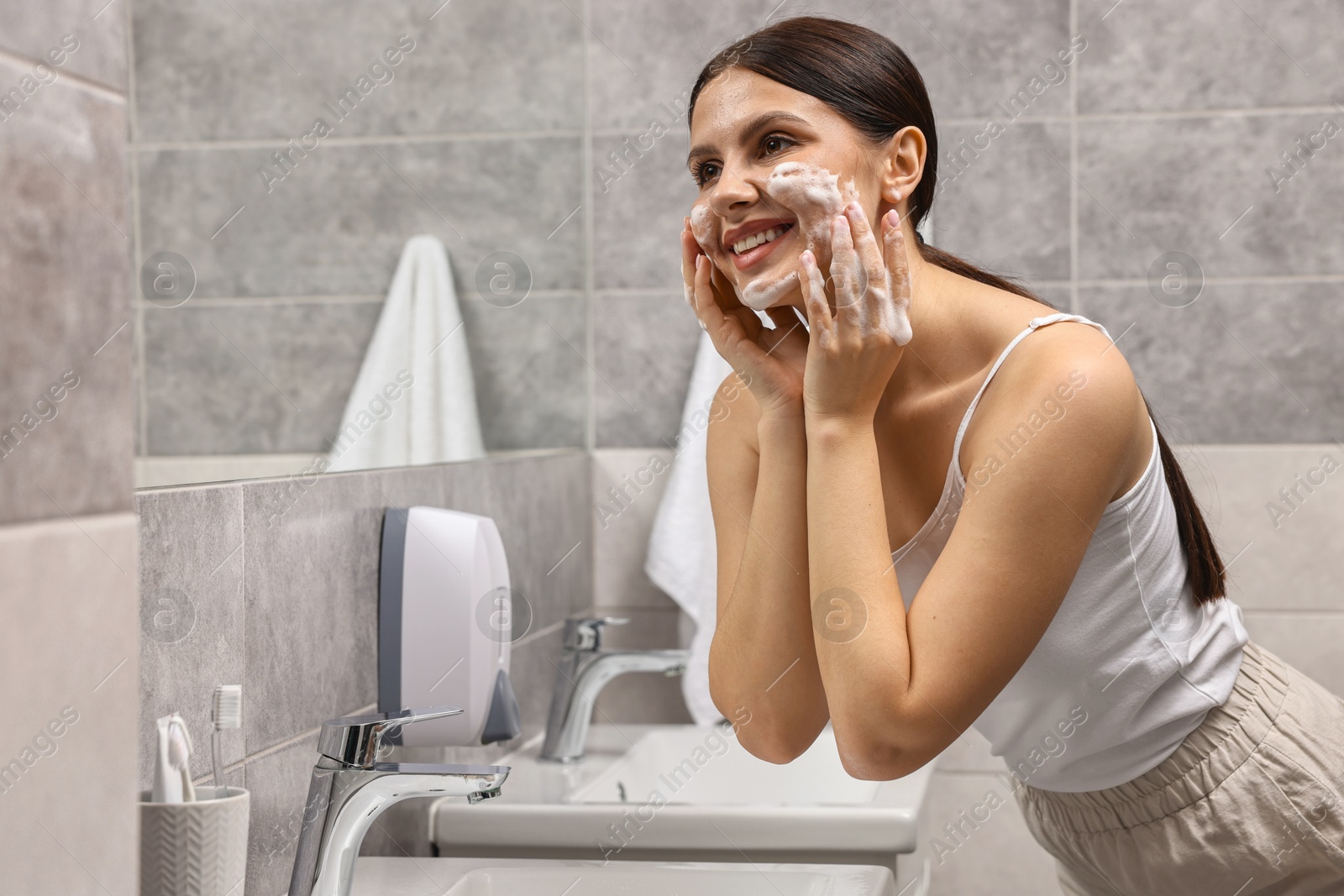 Photo of Woman washing her face with cleansing foam in bathroom