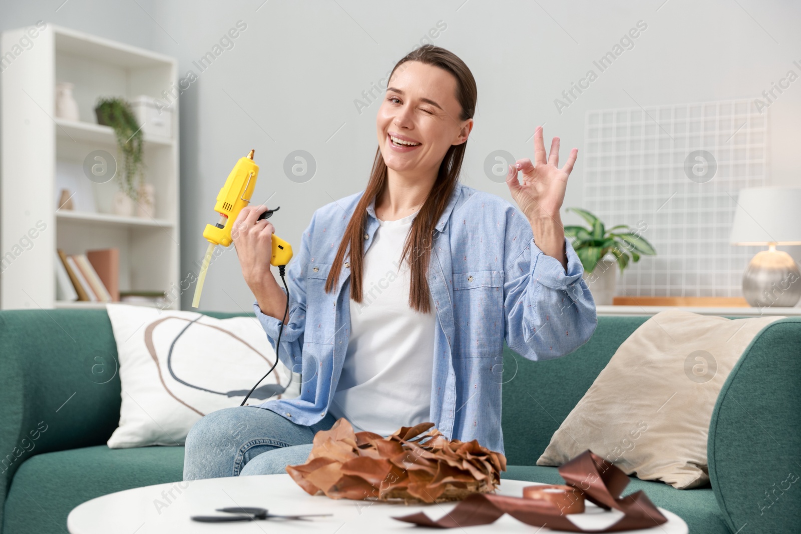 Photo of Woman with hot glue gun showing OK gesture at table indoors