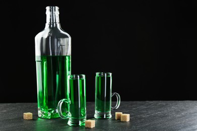 Photo of Delicious absinthe in shot glasses, bottle and brown sugar cubes on grey textured table against black background. Space for text