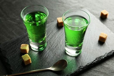 Photo of Delicious absinthe in shot glasses, spoon and brown sugar cubes on dark textured table, closeup