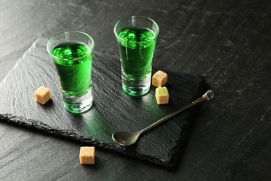 Photo of Delicious absinthe in shot glasses, spoon and brown sugar cubes on dark textured table, closeup