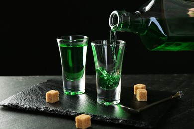 Photo of Pouring delicious absinthe from bottle into shot glass at grey textured table against black background, closeup