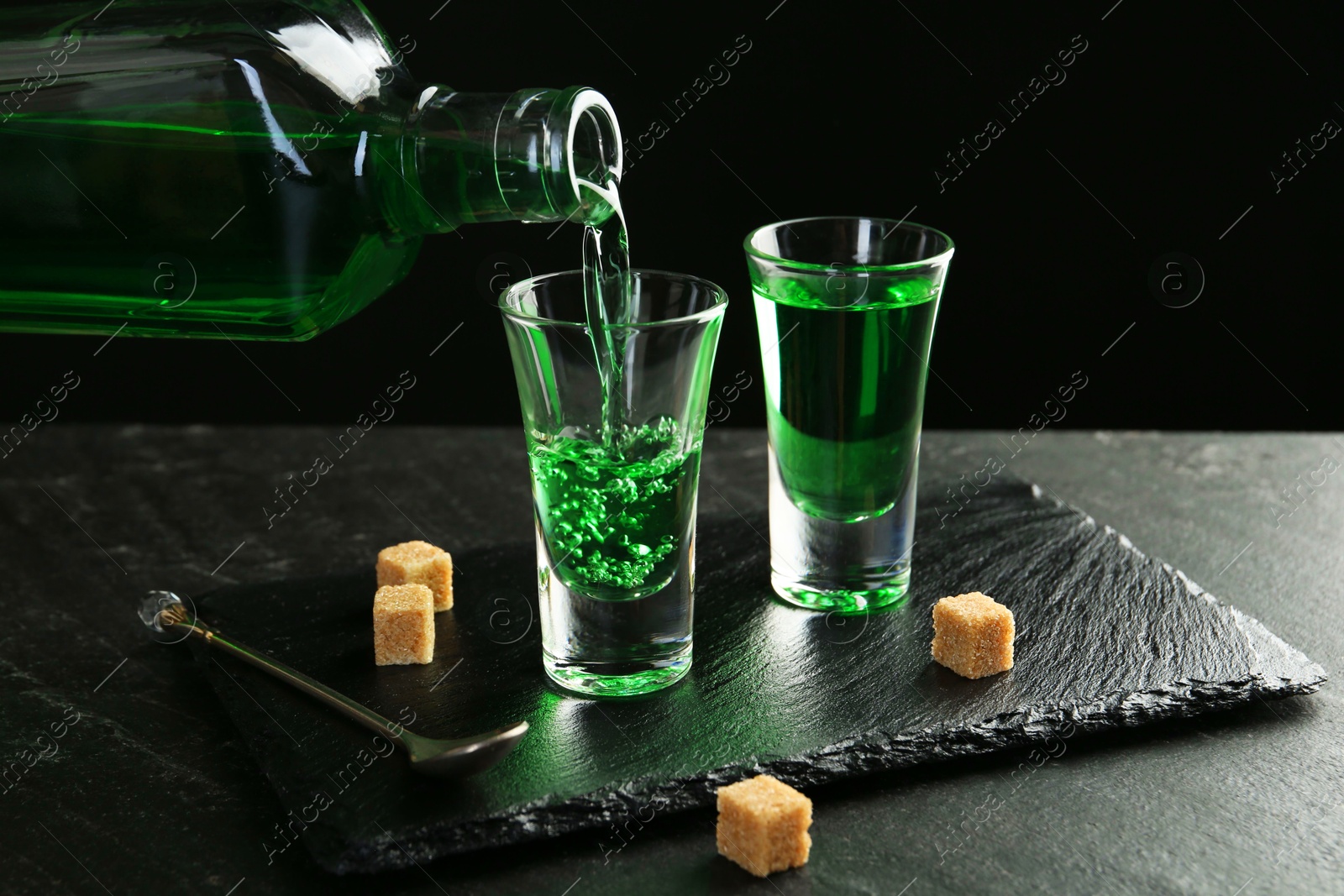 Photo of Pouring delicious absinthe from bottle into shot glass at grey textured table against black background, closeup