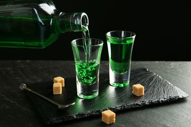 Photo of Pouring delicious absinthe from bottle into shot glass at grey textured table against black background, closeup