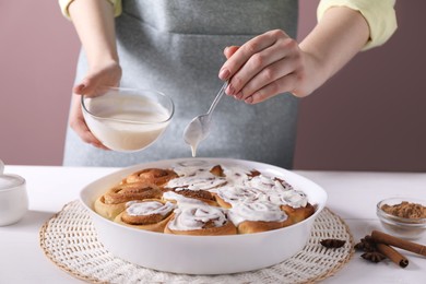 Photo of Woman spreading frosting onto freshly baked cinnamon rolls at white table against pink background, closeup