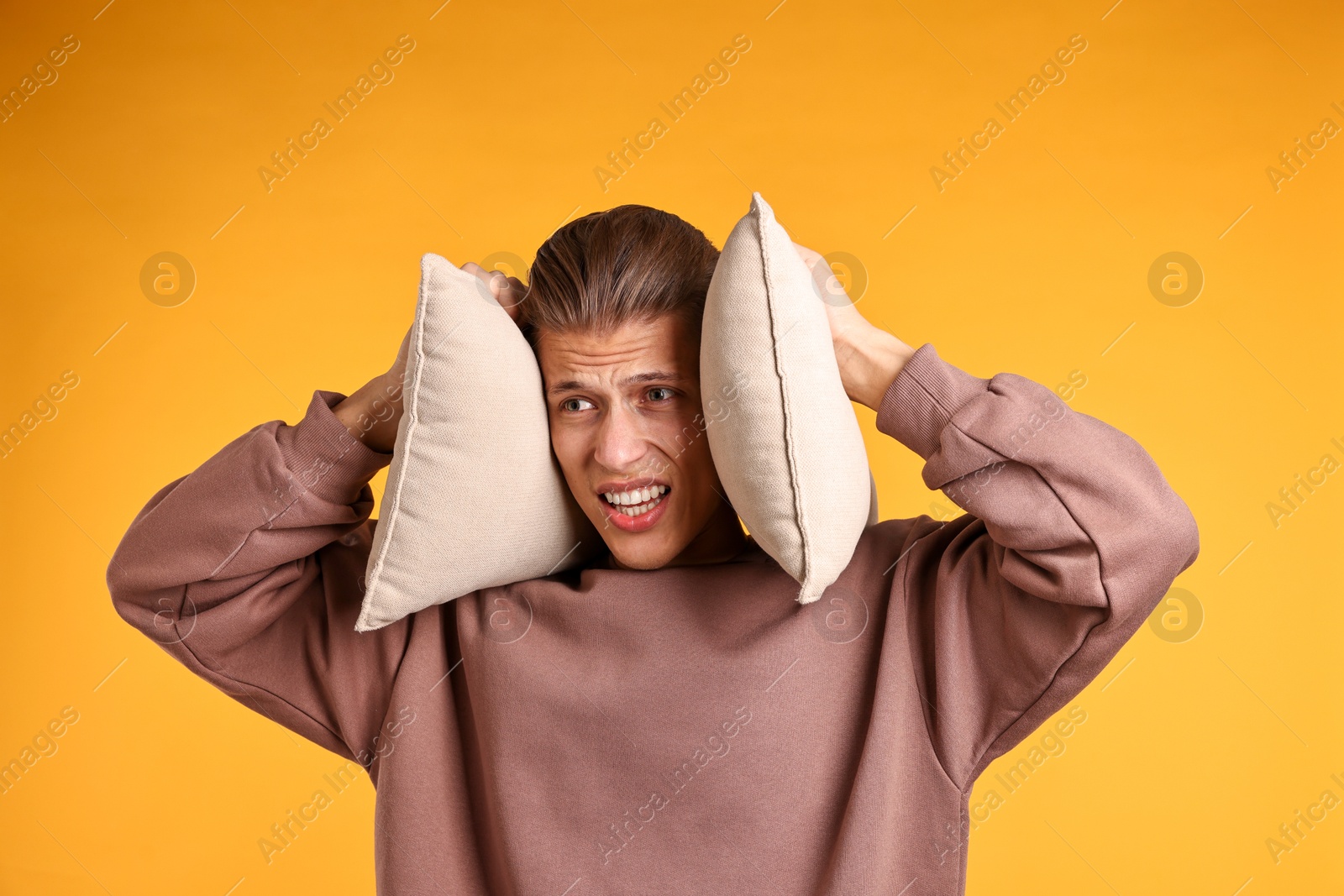 Photo of Annoyed young man covering his ears with pillows due to loud sound on orange background