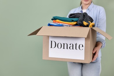 Photo of Woman holding donation box with clothes on pale olive background, closeup. Space for text