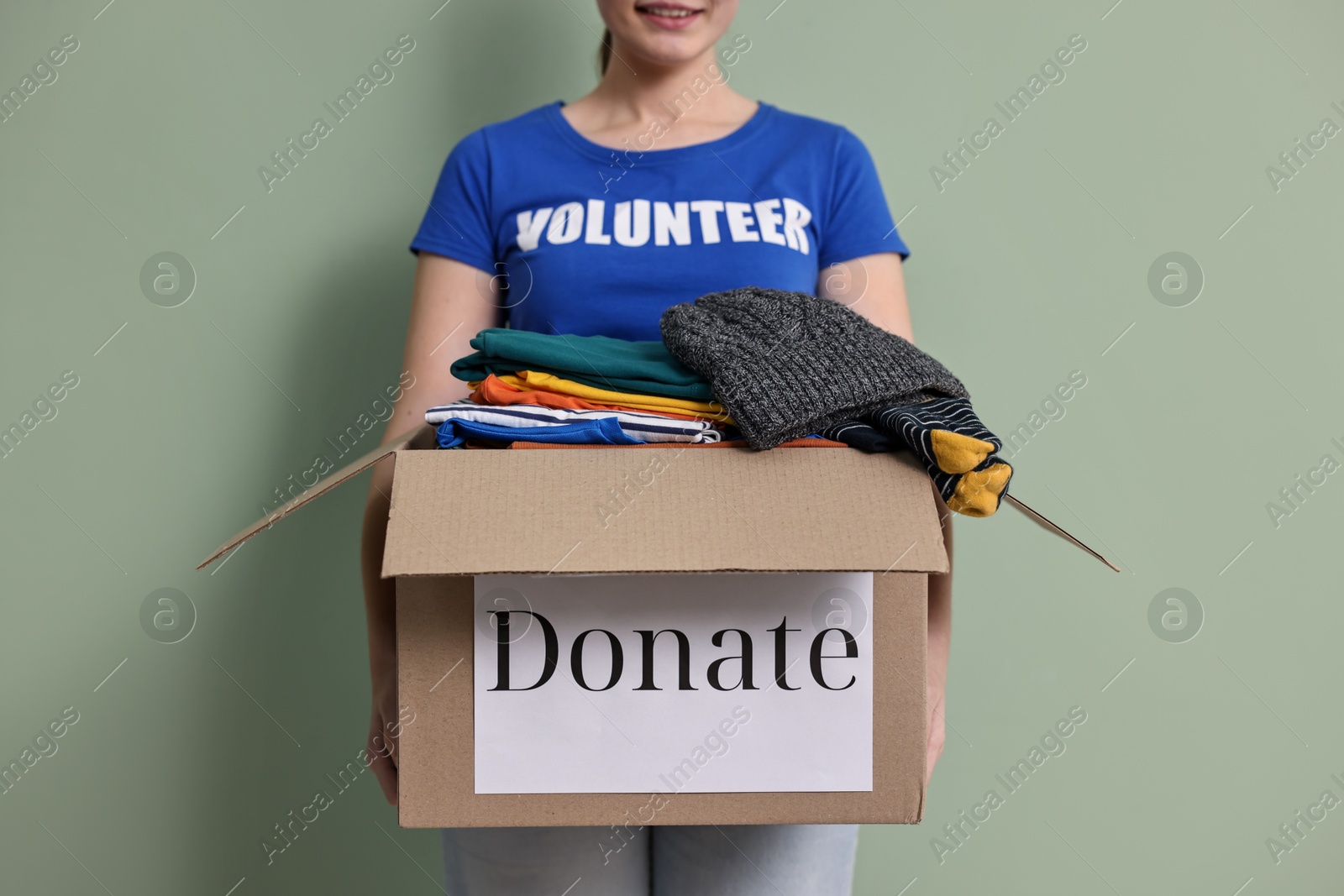 Photo of Woman holding donation box with clothes on pale olive background, closeup