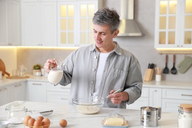 Photo of Making dough. Man pouring milk into bowl at white marble table in kitchen