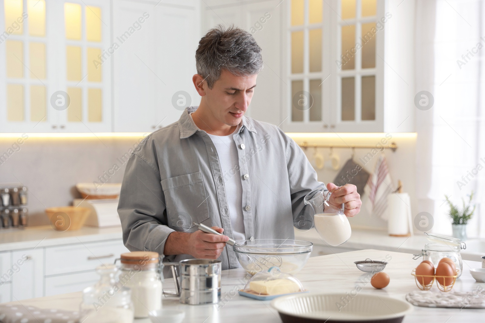 Photo of Making dough. Man pouring milk into bowl at white marble table in kitchen