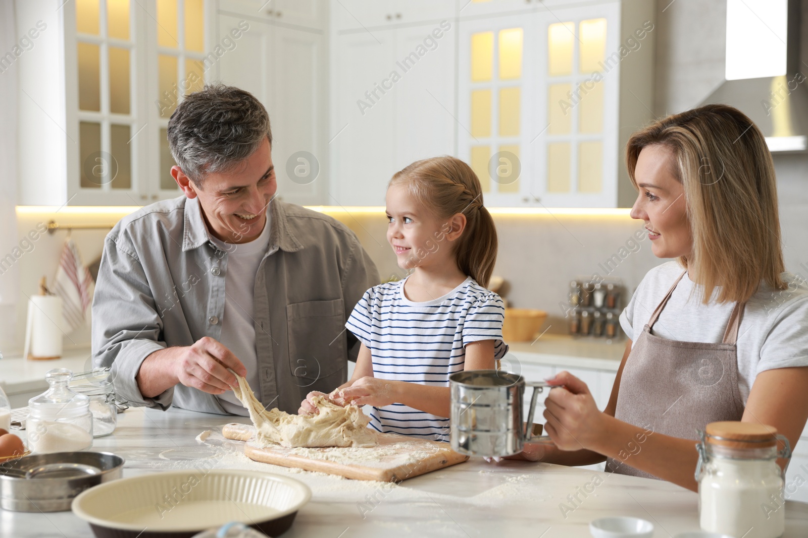 Photo of Happy parents and their daughter making dough at white marble table in kitchen