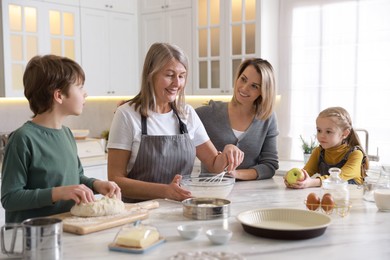 Little kids with their mother and grandmother making dough at white marble table in kitchen